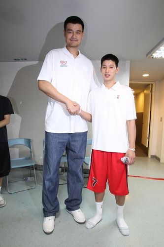 A very tall man in casual clothes shaking hands and taking a picture with a teenage boy in basketball shorts.